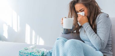 Someone blowing their nose into a tissue, holding a cup of tea.