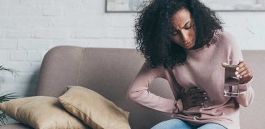 A woman sitting on a couch, holding a glass of water and clutching her stomach